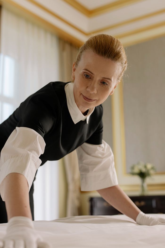 about-img-01 A housekeeper in uniform skillfully arranges bed linen in a luxurious hotel room.