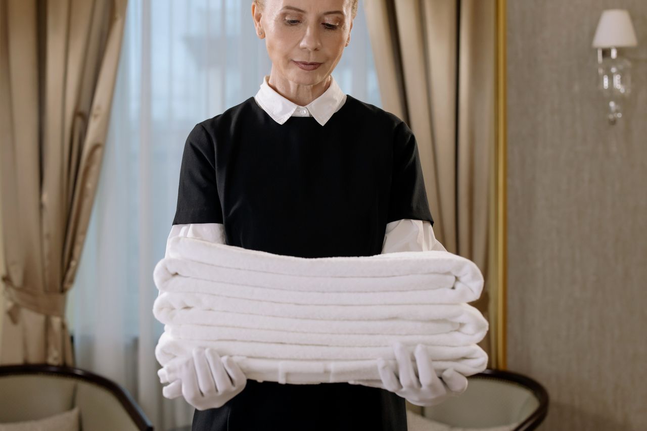gallery-02 Professional housekeeper holding a stack of fresh towels in a luxurious hotel room, showcasing elegance and service.