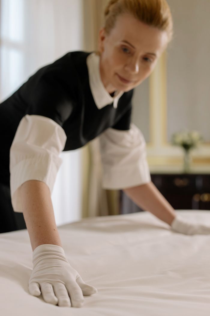 services-img A housekeeper elegantly arranges bed sheets in a luxurious hotel room, showcasing hospitality expertise.